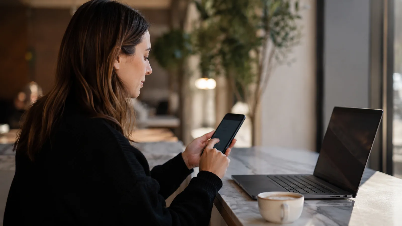 A woman sitting at a cafe counter checking her phone, with a laptop and a cup of coffee on the marble table beside her