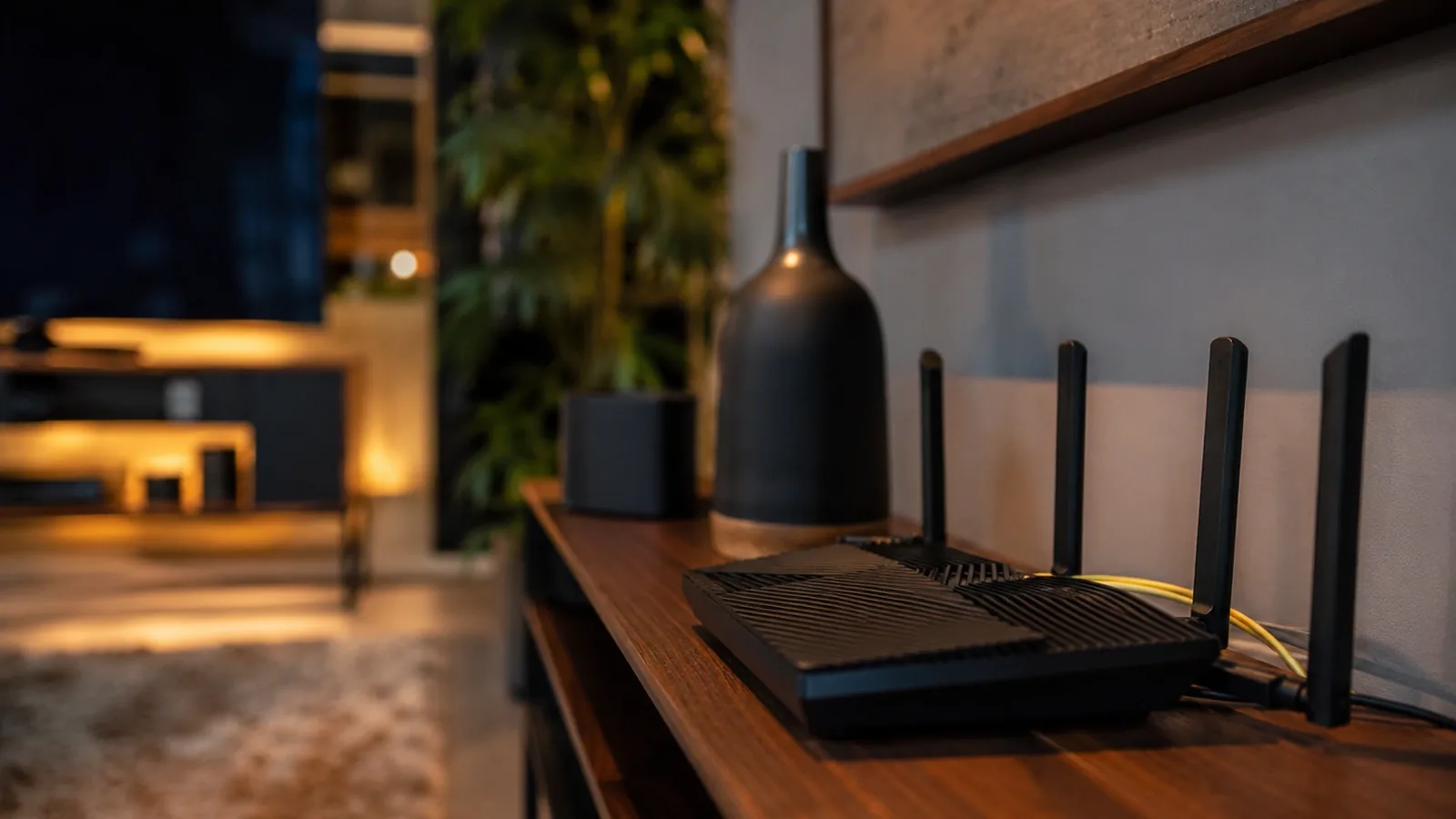 A close-up of a black Wi-Fi router with four antennas on a wooden shelf, a living room and television out of focus behind it