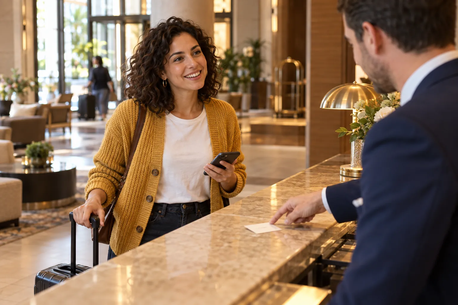 A woman in a mustard cardigan with her suitcase at a hotel reception desk asks the receptionist for the Wi-Fi network name