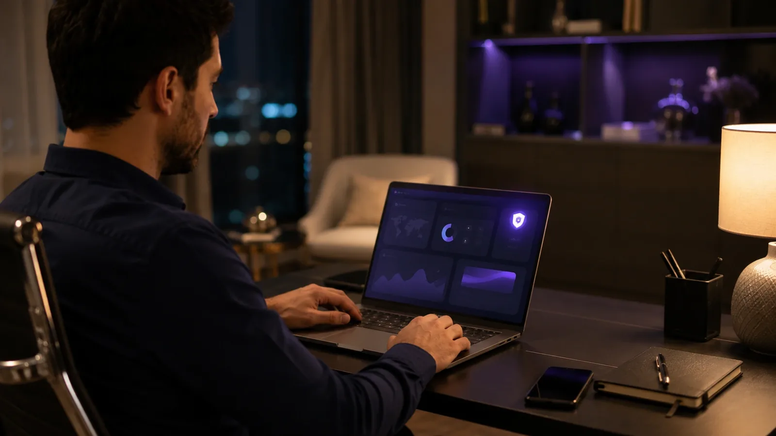 A man working late at a home desk, a laptop in front of him showing a dashboard with charts and a security shield icon